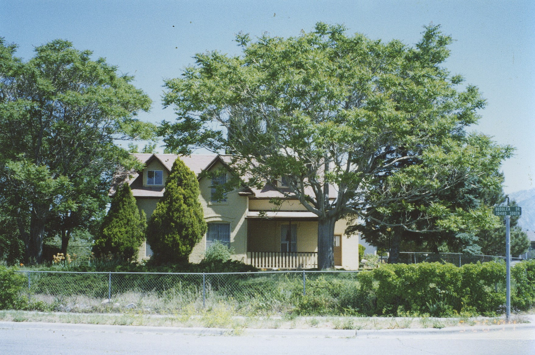 A yellow house with a small porch, surrounded by trees and a chain-link fence. Street sign visible at the corner.