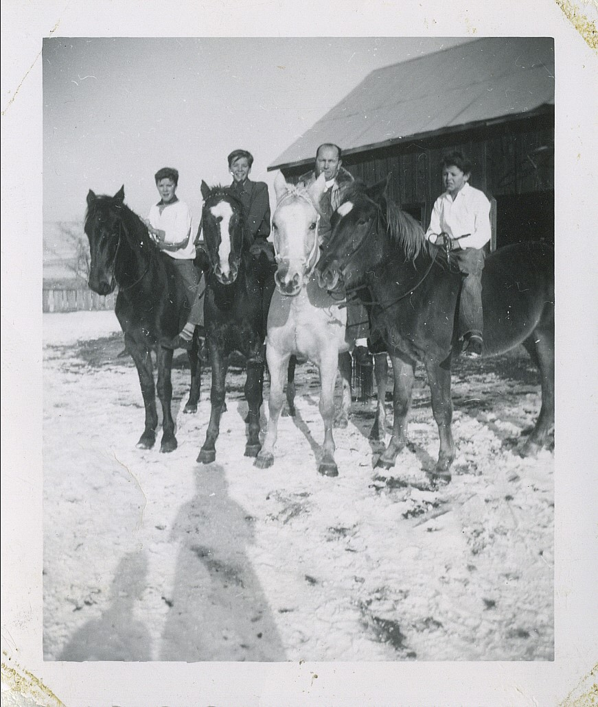 Four people riding horses in a snowy area, with a barn in the background.