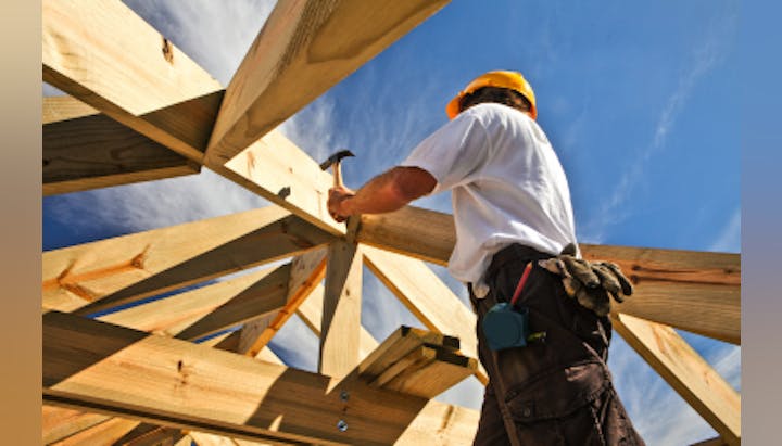 A construction worker wearing a hard hat is hammering nails into a wooden roof frame under a clear blue sky.