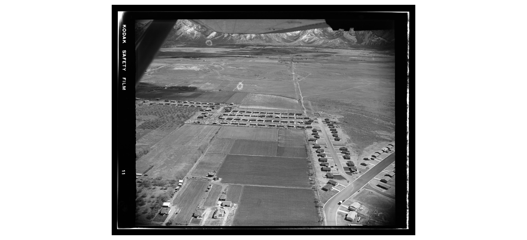 Aerial view of a grid-like residential area, roads, and fields with mountains in the background.