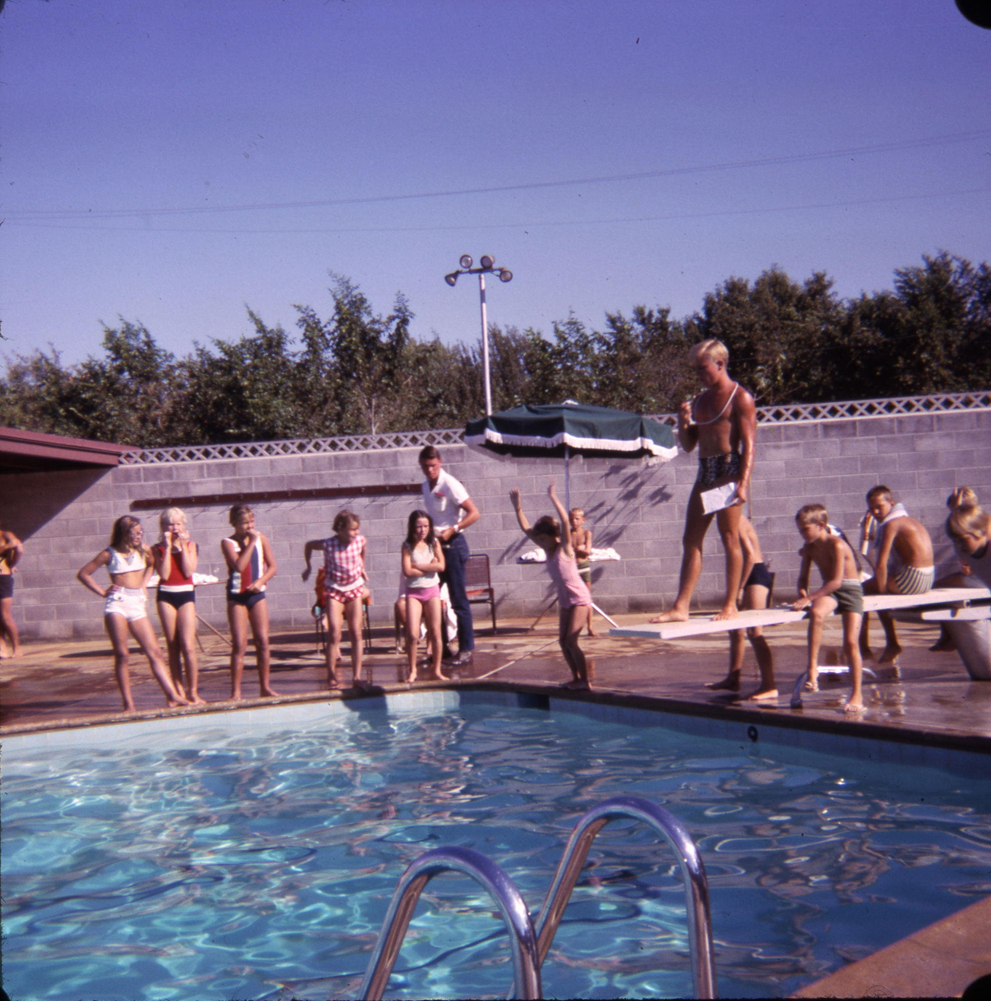 A group of children at a pool, some standing on a diving board, others watching by the poolside.