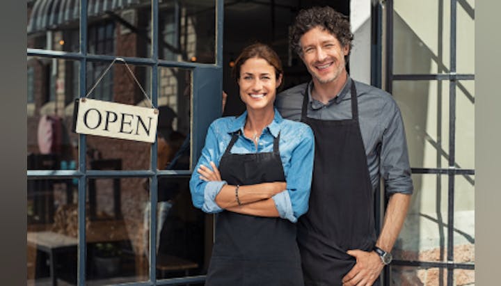 Two people in aprons smiling outside a shop with an "OPEN" sign.