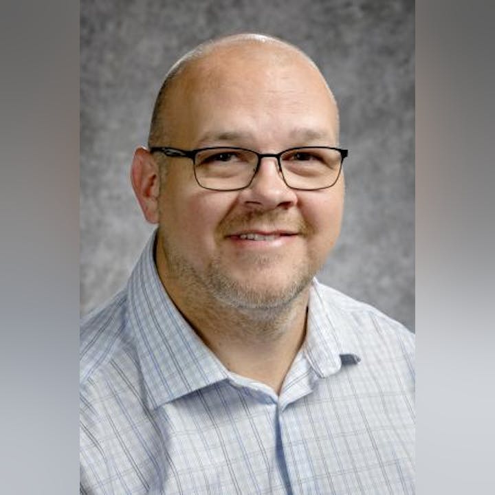 A person with glasses wearing a checked shirt, smiling against a gray background.