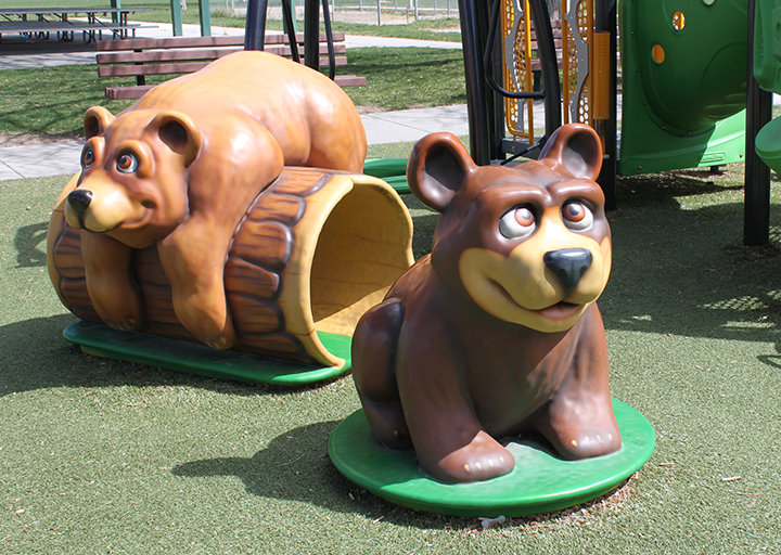 Two playful bear statues in a playground setting, one on a log, the other sitting on the ground.
