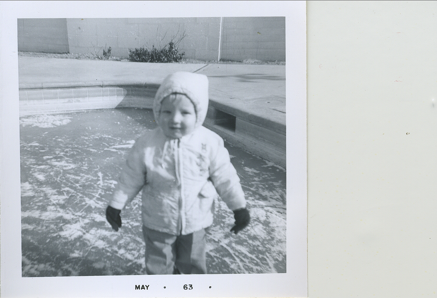 A child bundled in winter clothes stands by a pool. The photo is dated May 1963.