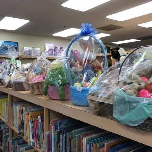 Gift baskets wrapped in plastic, filled with toys and plush items, are displayed on a bookshelf in a library.