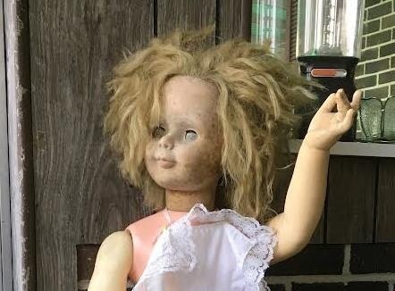 A vintage doll with disheveled hair, wearing a white dress, and posed with one arm raised in a rustic setting.