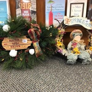 Two decorative wreaths, one rustic with pinecones, the other spring-themed with a scarecrow. Books are displayed in the background.
