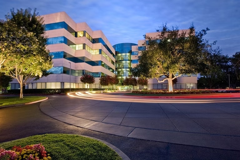 Modern office building at dusk with light trails from cars, surrounded by trees and landscaping.