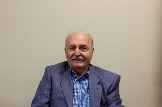 An elderly man wearing a blue checkered shirt and a blazer against a plain background.