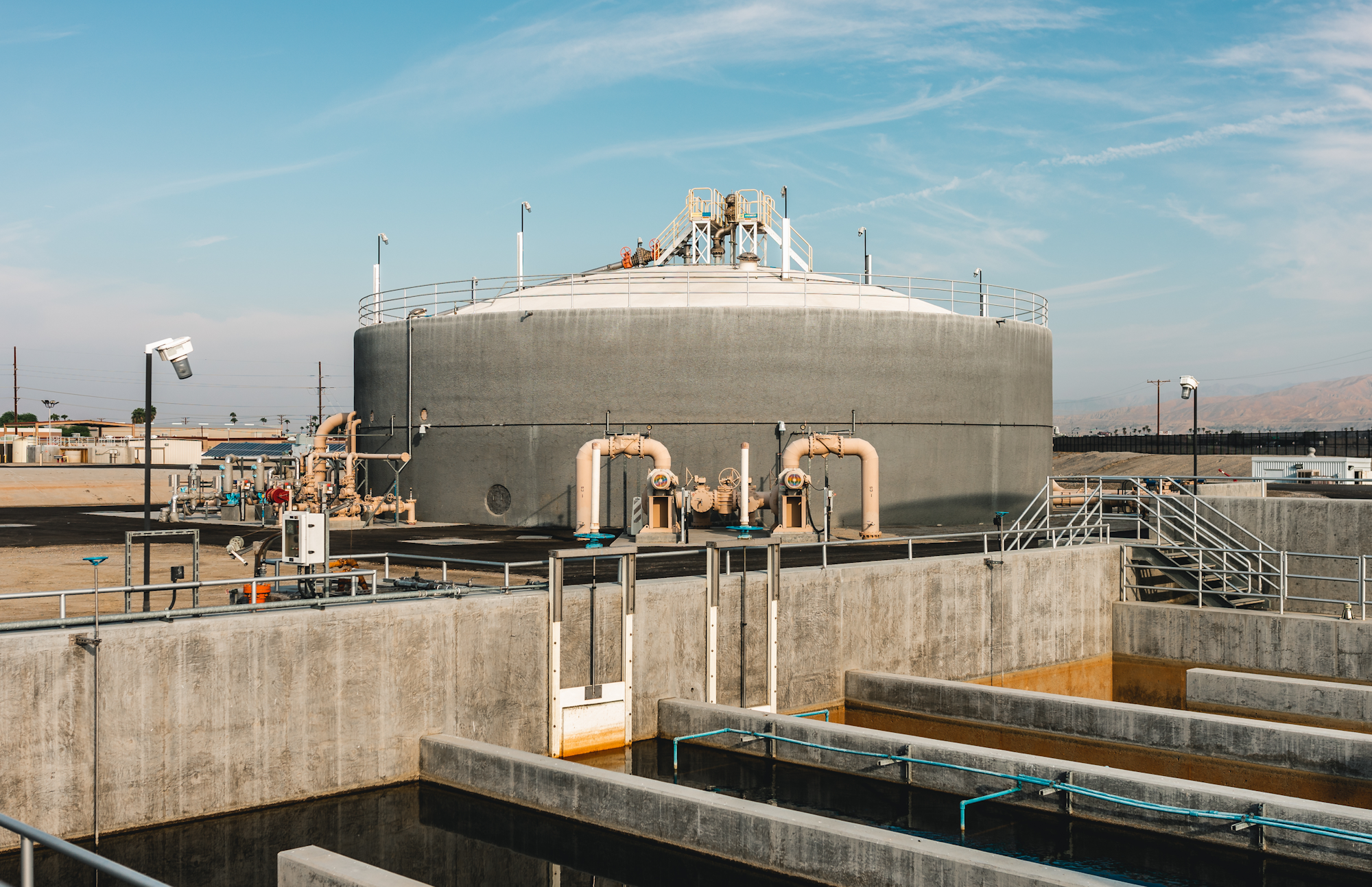 The image shows a large industrial facility with a cylindrical tank, pipes, and concrete structures under a clear blue sky.