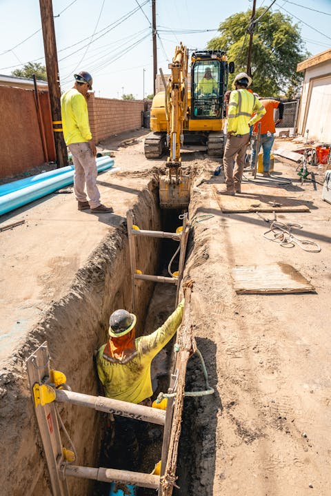 Construction workers excavating a trench with an excavator; one worker is inside the trench with safety gear.