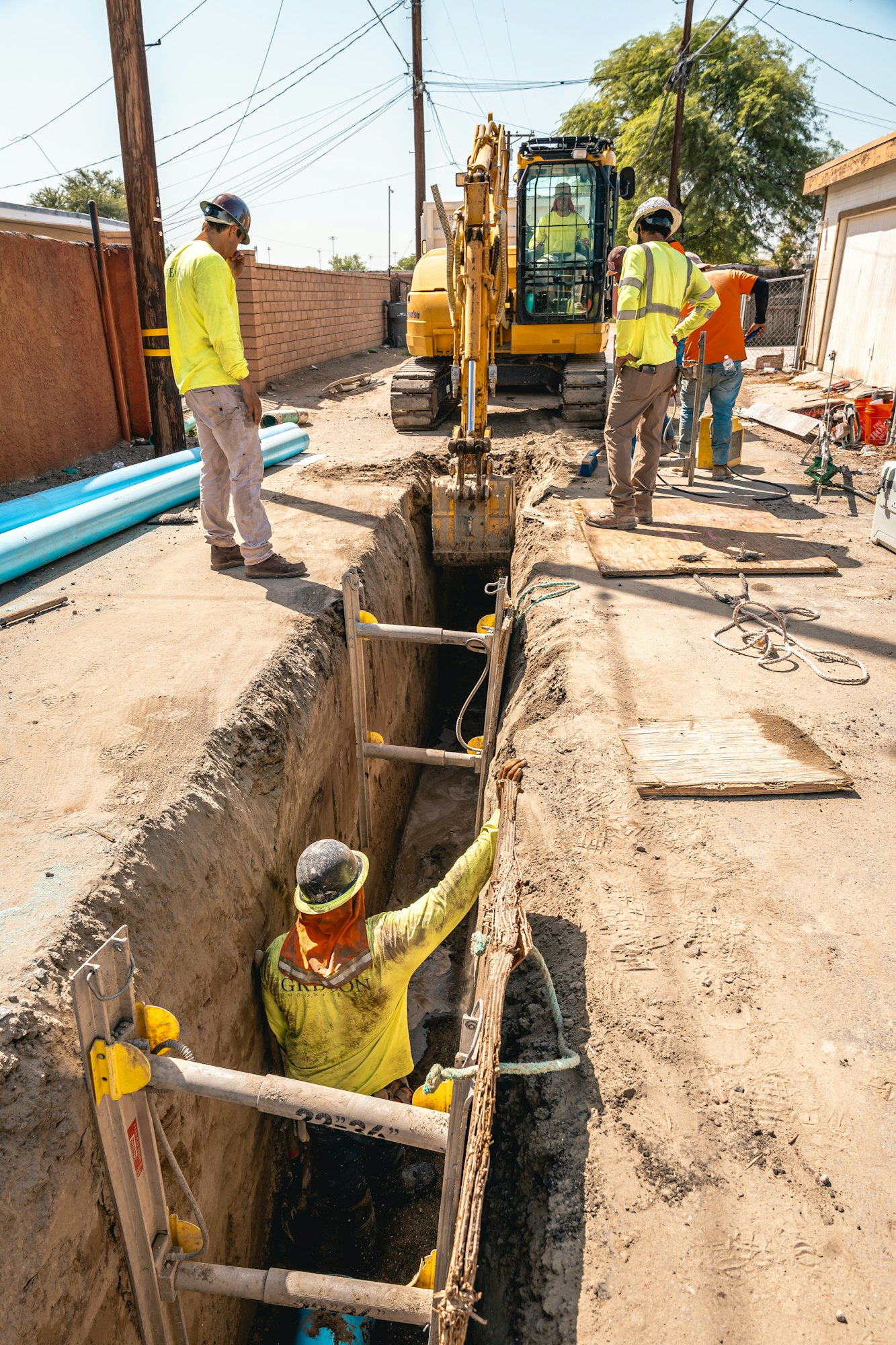 Construction workers excavating a trench with an excavator; one worker is inside the trench with safety gear.