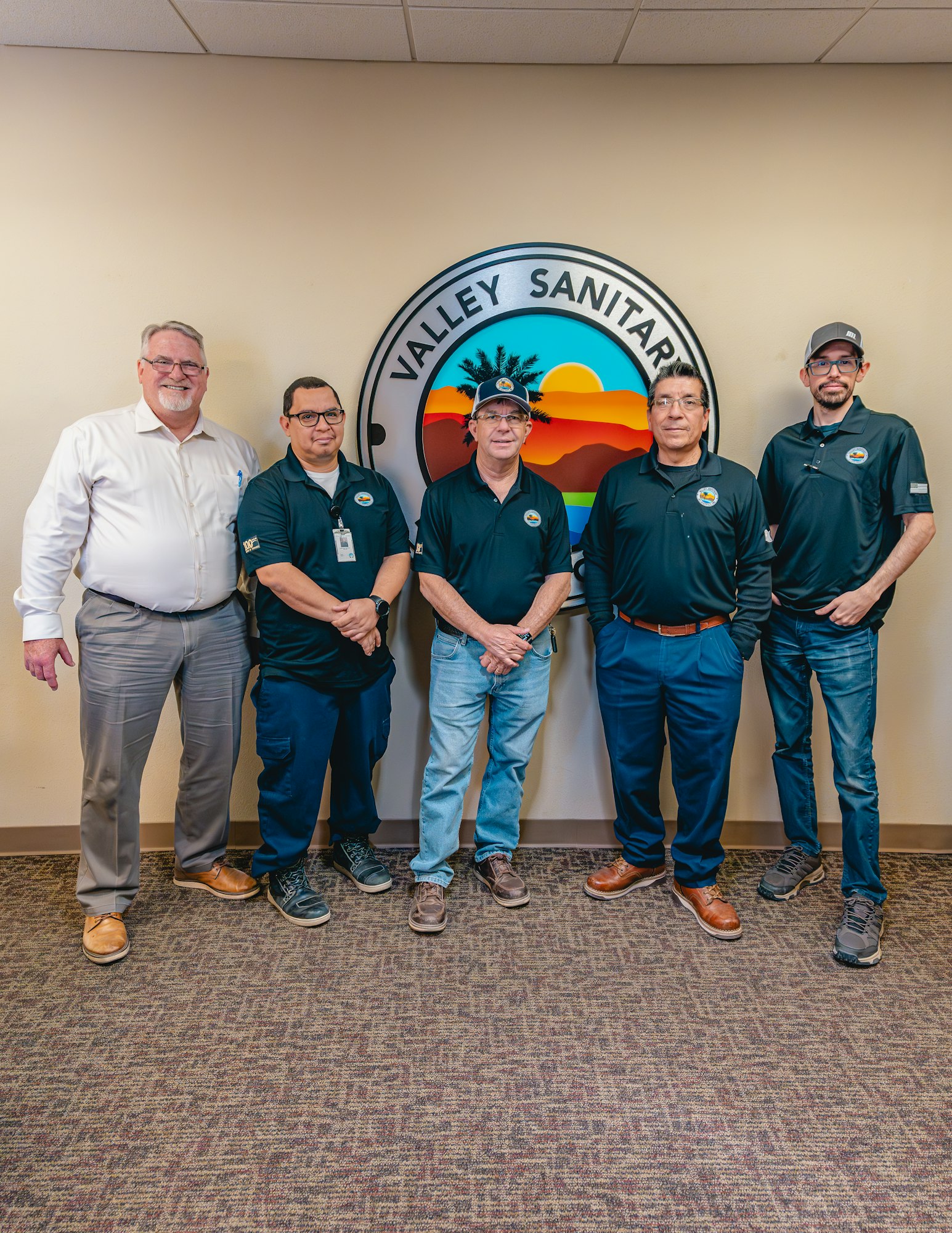 Five men standing in front of a "Valley Sanitary" sign, wearing casual and work attire.