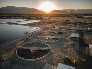 Circular treatment tanks at a water facility with sunrise, mountains, and buildings in the background.