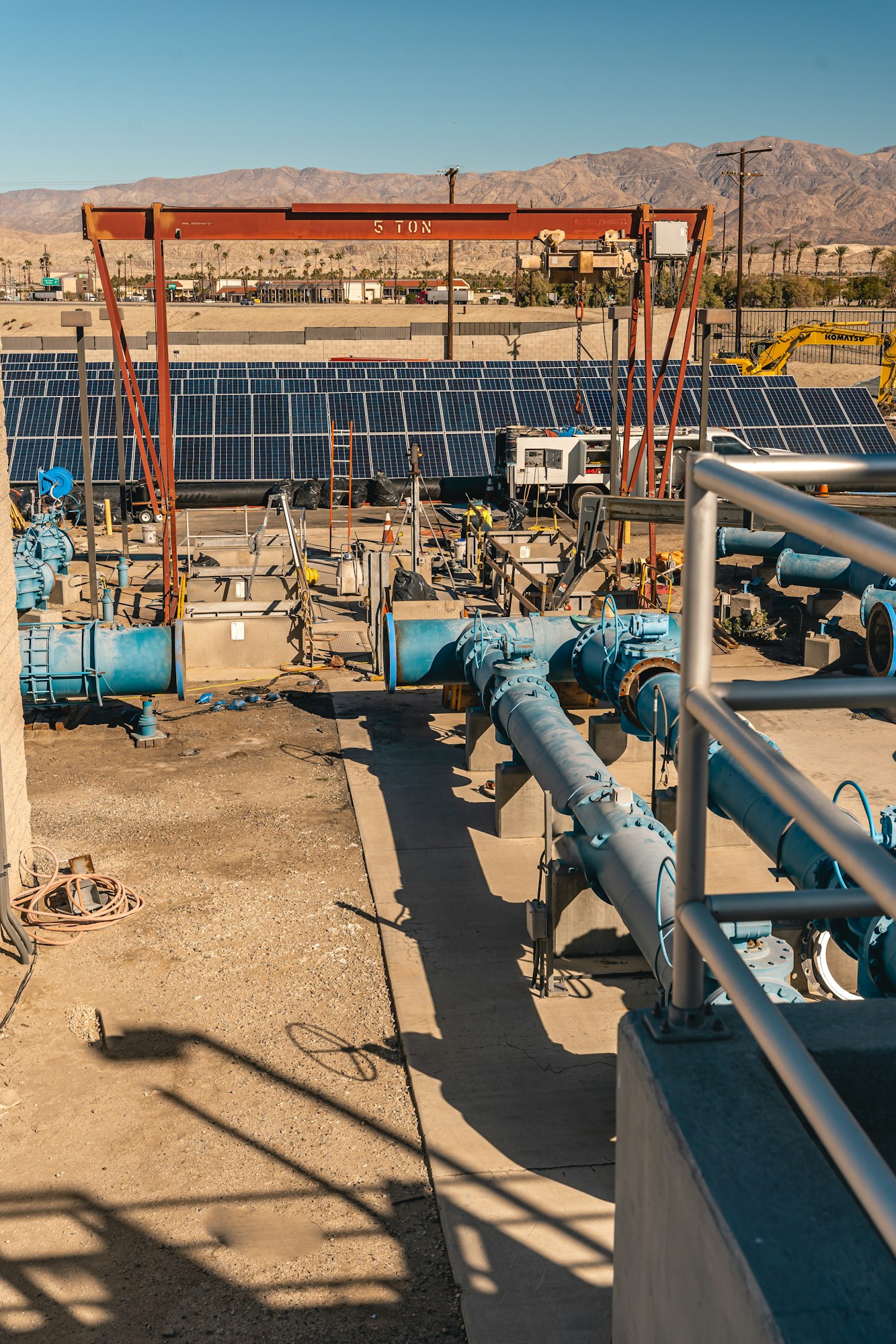 Industrial site with blue pipes and solar panels under a clear sky, mountains in the background.