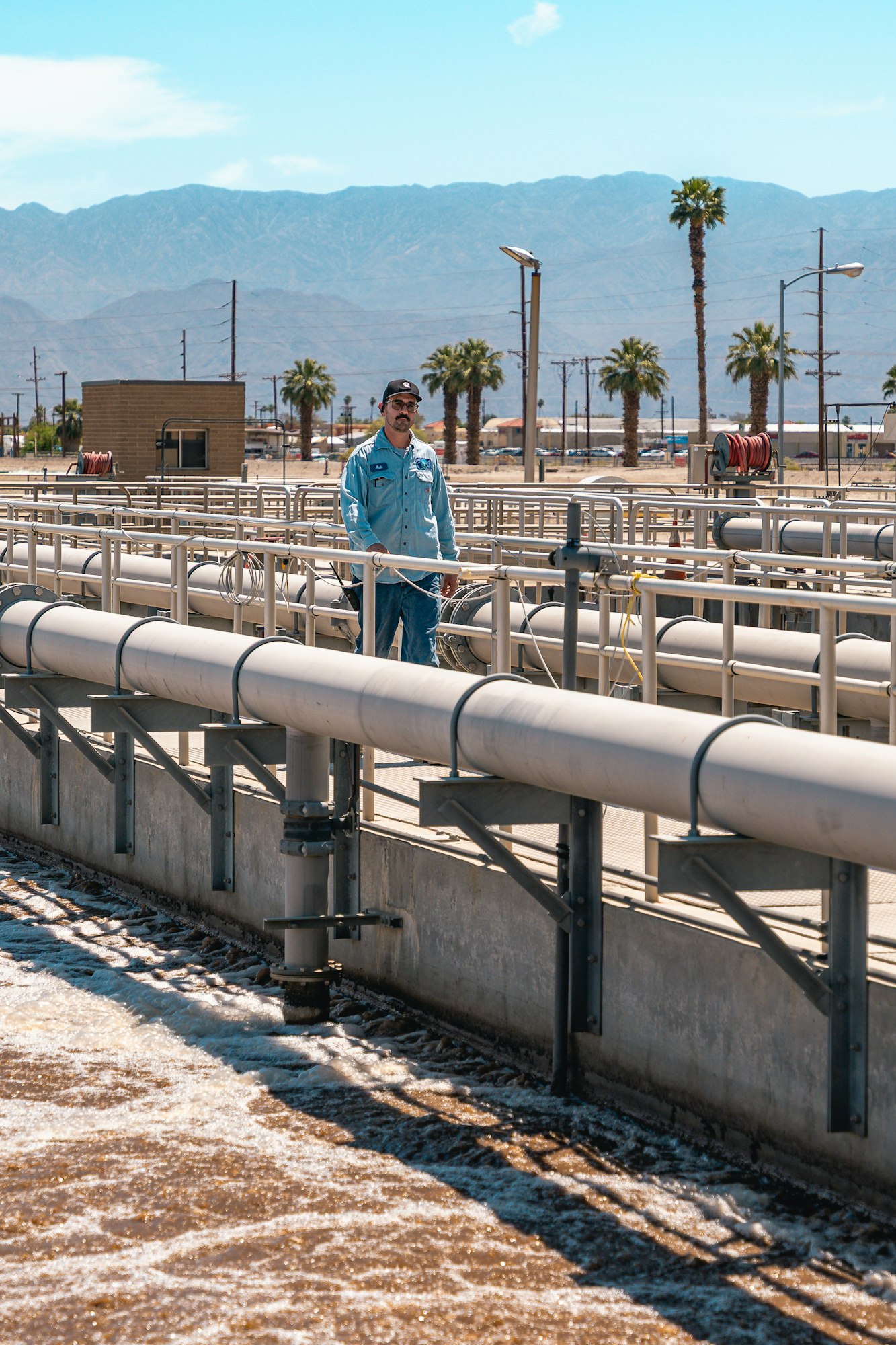A person in a uniform stands at an industrial facility with pipes, near mountains and palm trees under a clear sky.