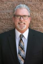 A man with glasses in a suit and patterned tie, standing against a brick wall.