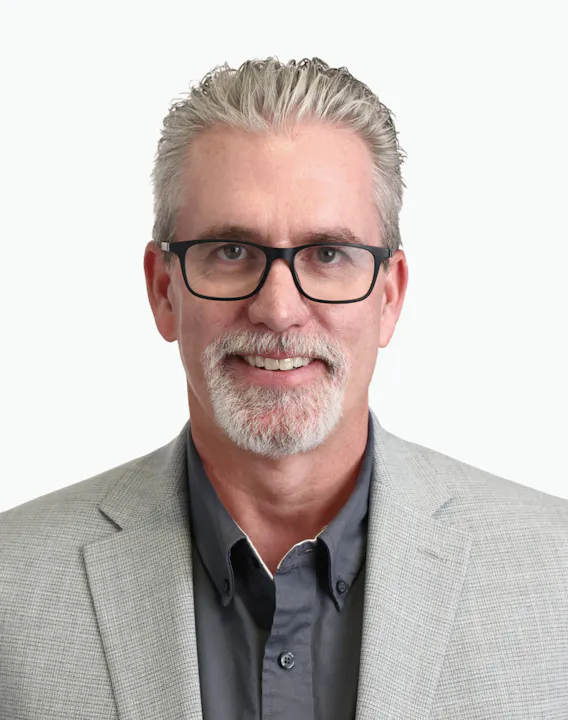 The image features a smiling man with glasses, wearing a gray blazer over a dark shirt. The background is plain and light-colored.