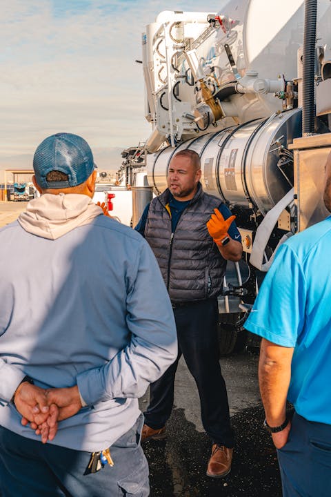 A man in an orange glove gestures while talking to two others near a large industrial vehicle.