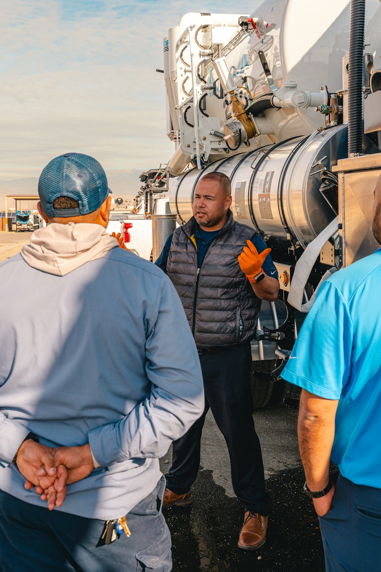 A man in an orange glove gestures while talking to two others near a large industrial vehicle.