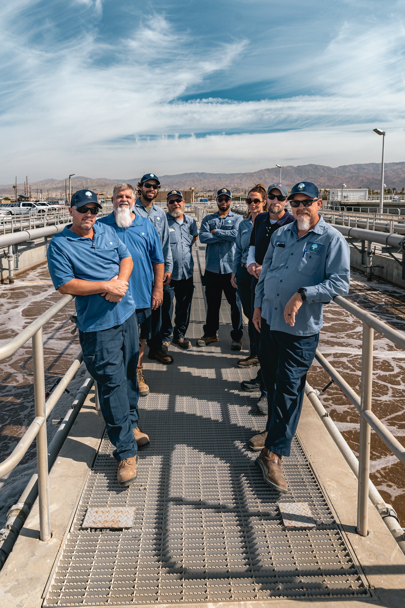 A group of people in blue uniforms standing on a facility walkway with mountains in the background.