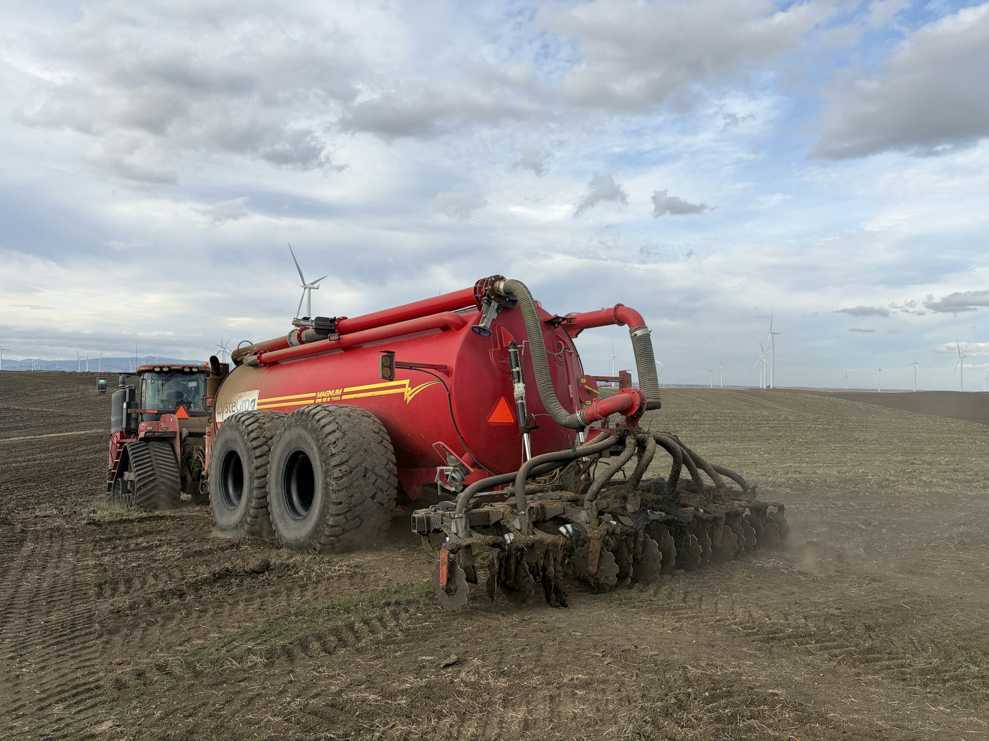 A red tractor pulls a large tank with equipment, working on a field with wind turbines visible in the background.
