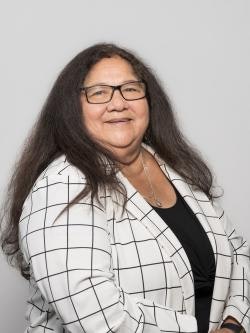 A person with long hair and glasses, wearing a checkered blazer over a black top, poses against a plain background.