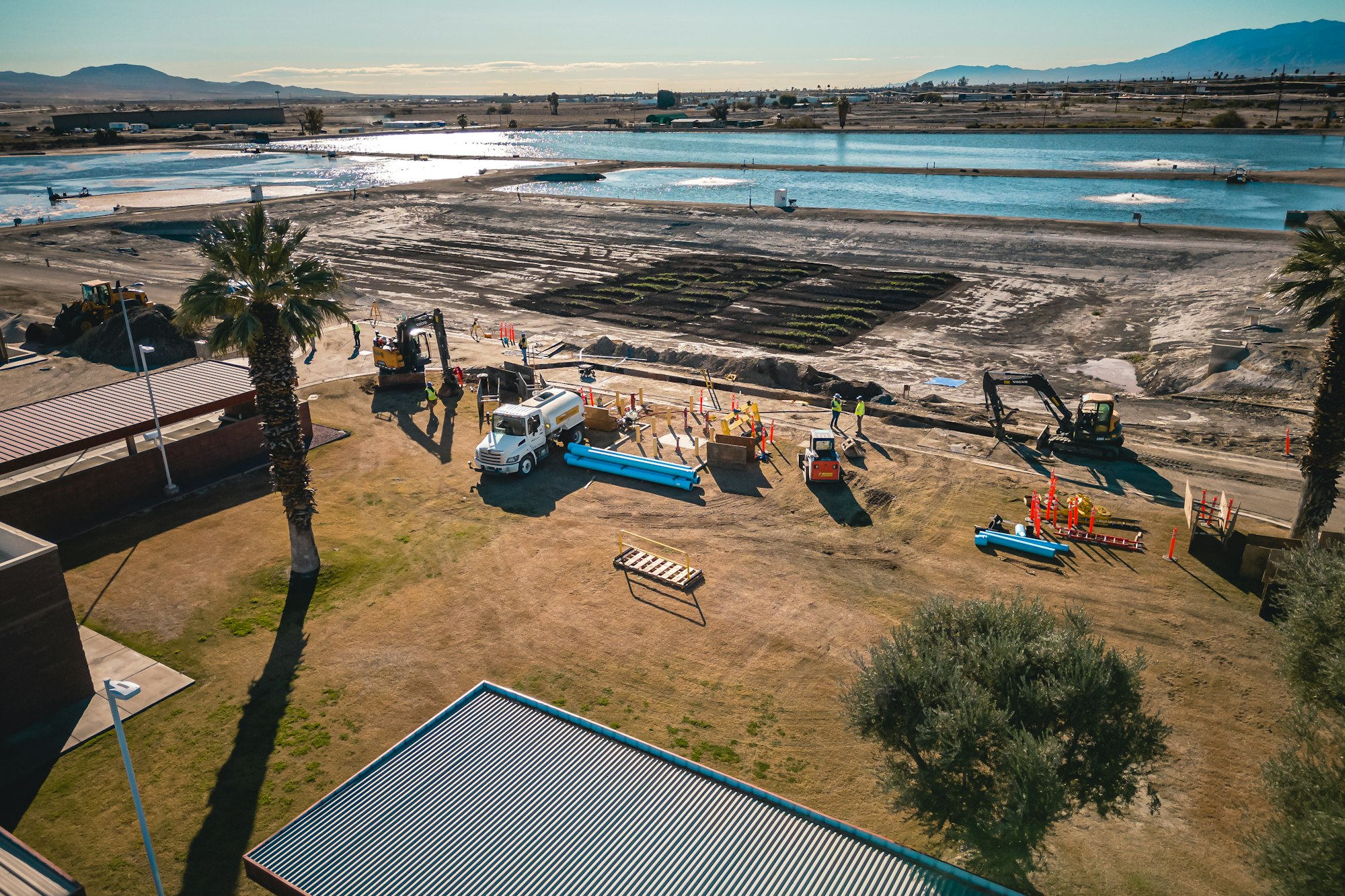 Construction site near water with vehicles, equipment, and workers; palm trees and landscape in the background.