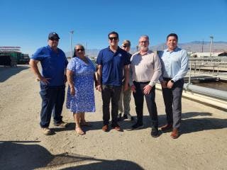 Six people standing outdoors on a sunny day, with pipes and a mountain in the background.