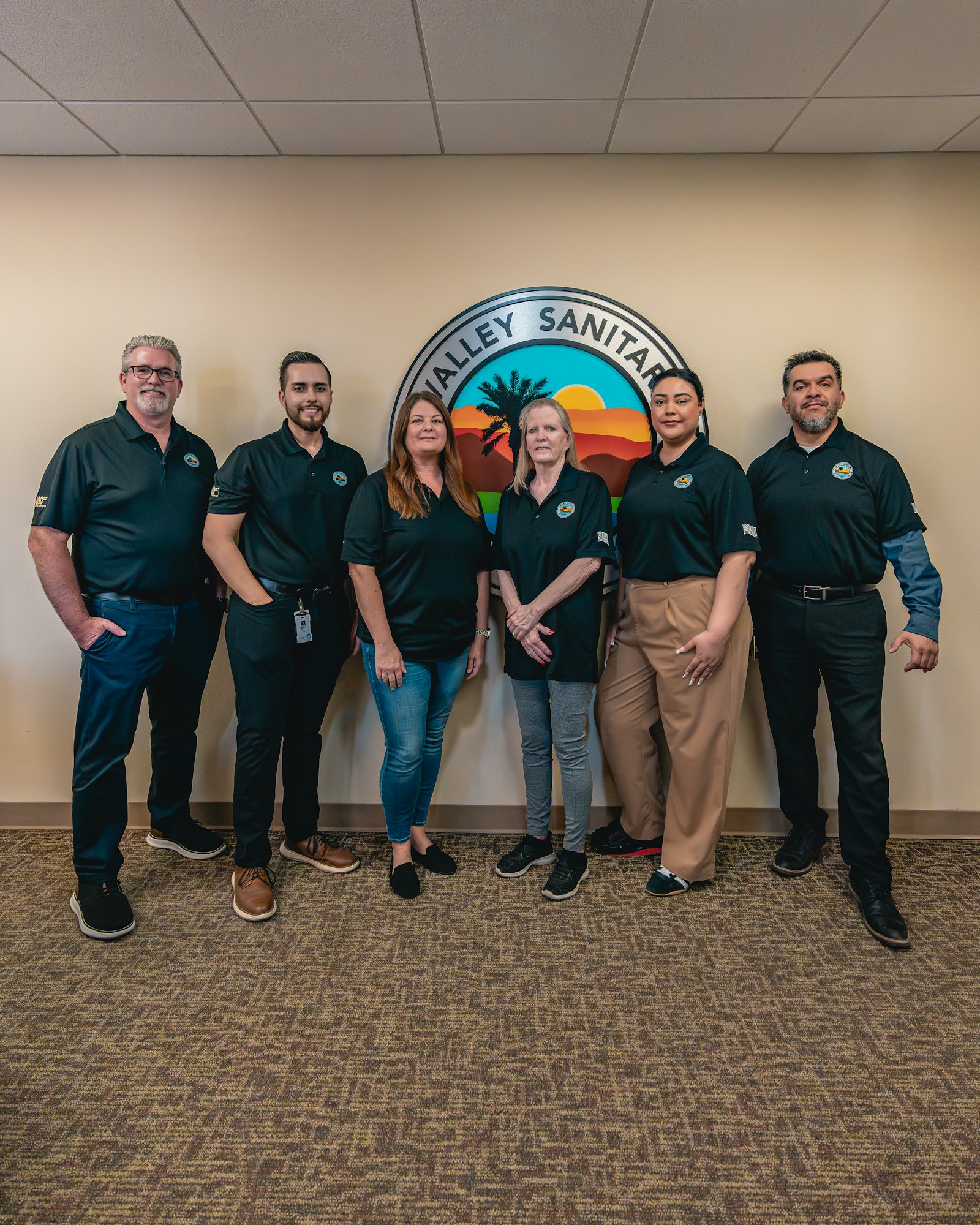A group of six people wearing black shirts standing in front of a "Valley Sanitary" logo on a beige wall.