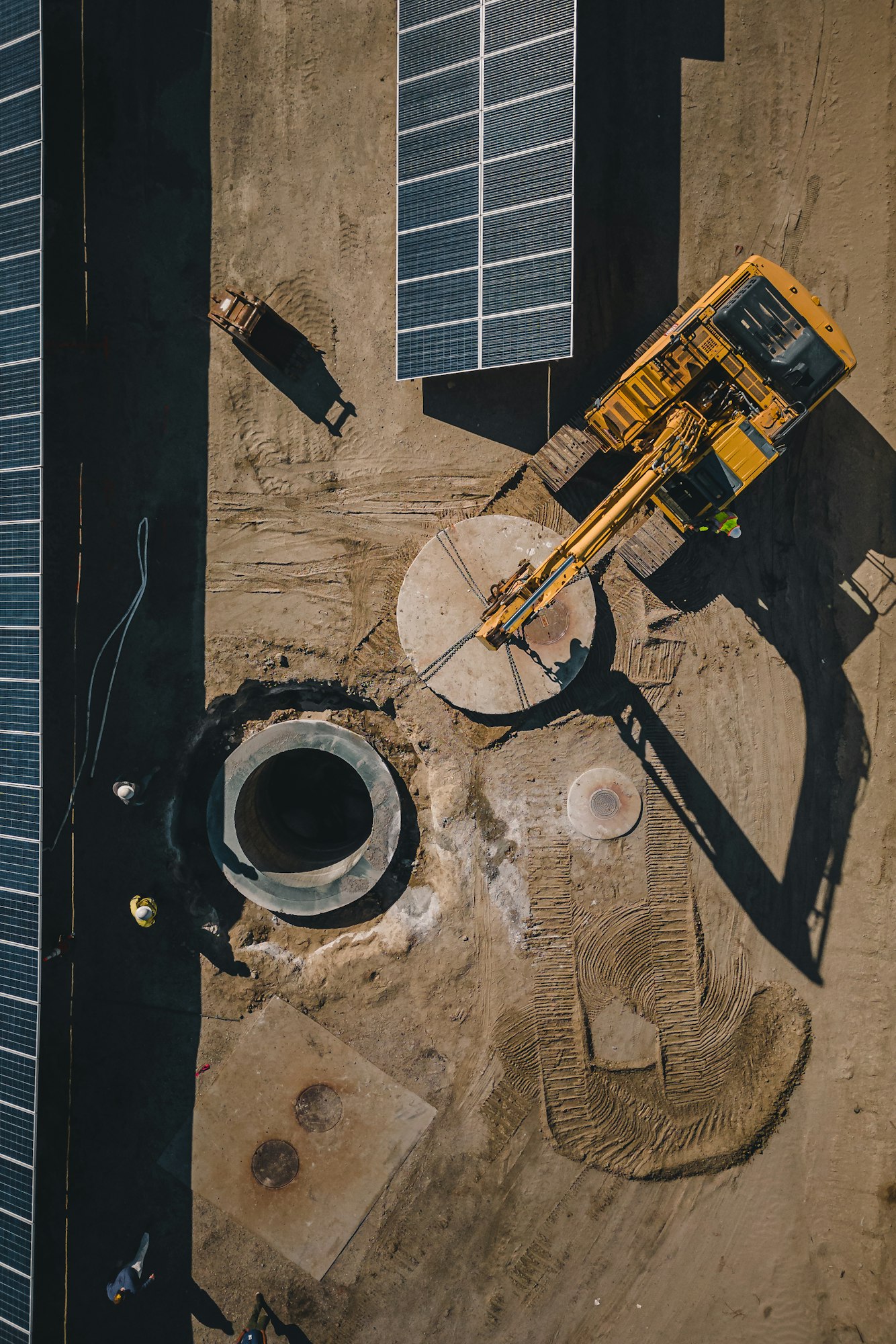 Construction site with solar panels, an excavator, and workers near a large circular structure.
