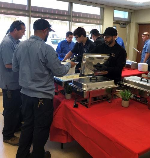 People serving and receiving food at a buffet with red tablecloths and chafing dishes.