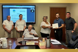 Meeting room scene with people standing, two holding certificates. There's a screen on the wall and a table with papers and a nameplate.