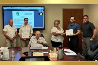 Meeting room scene with people standing, two holding certificates. There's a screen on the wall and a table with papers and a nameplate.