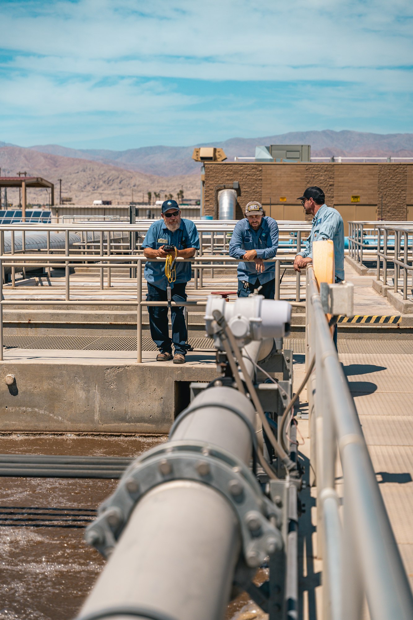 Three people in work attire standing on a platform at an industrial facility with mountains in the background.