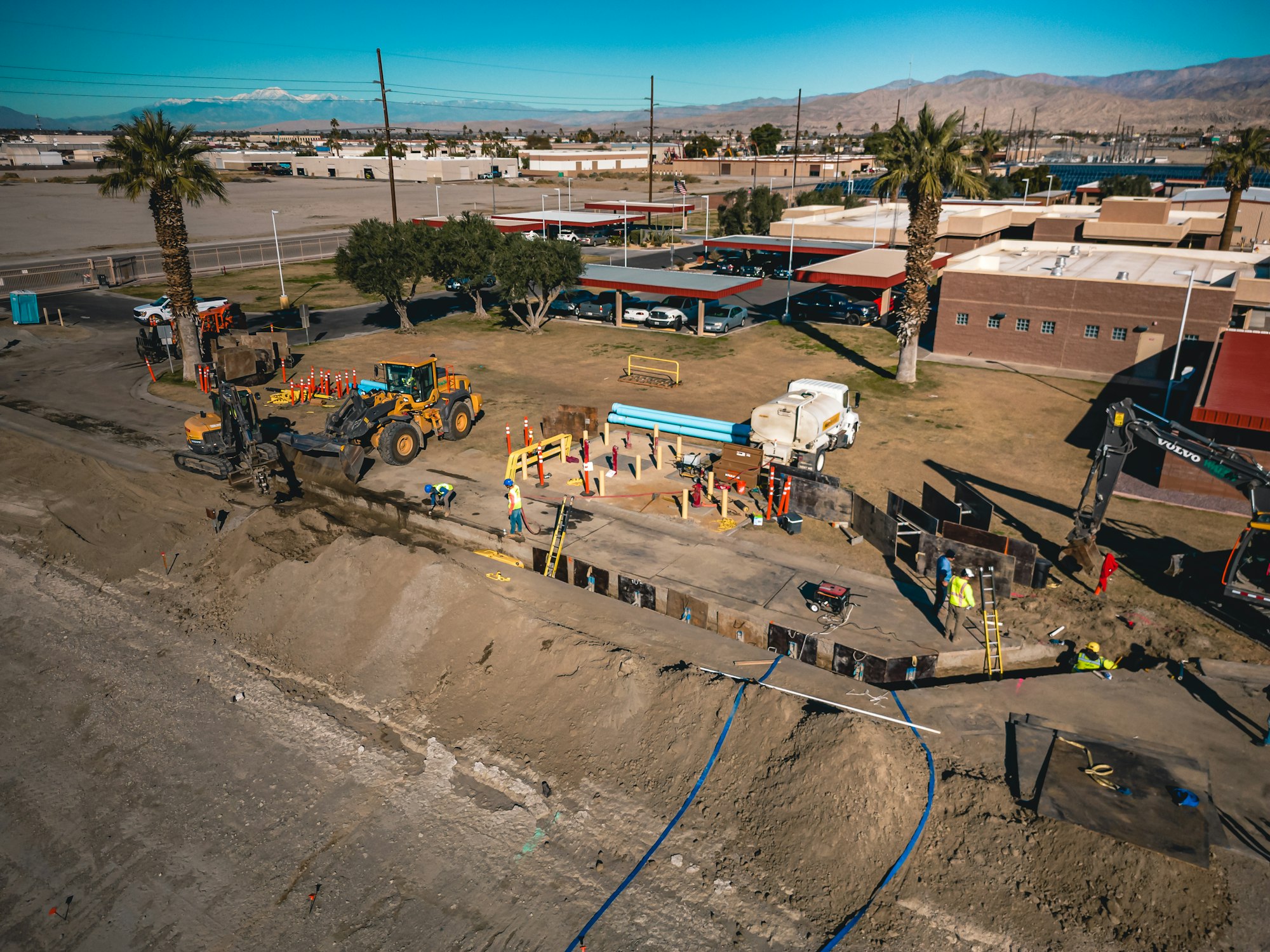 Construction site with heavy machinery, workers in safety gear, and equipment. Desert landscape and suburban area in the background.