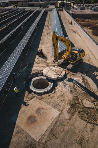 Construction site with an excavator lifting a circular concrete slab. Worker nearby, surrounded by solar panels and paved areas.