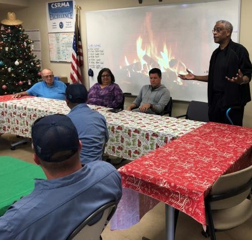 A group meeting in a room with holiday decorations, a Christmas tree, and a virtual fireplace on the screen.