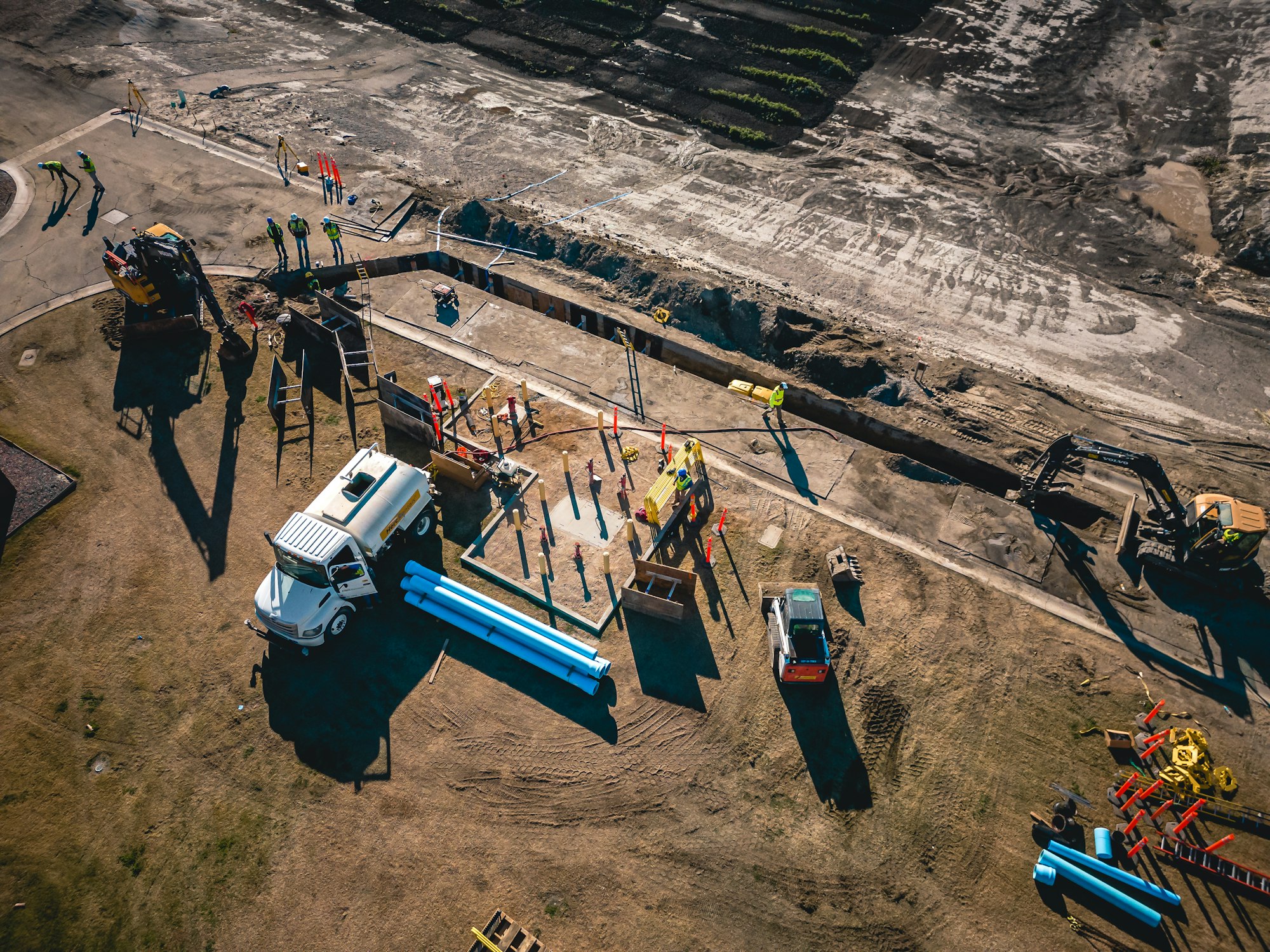 Construction site with vehicles, pipes, workers in safety vests, and excavators near a dirt trench.