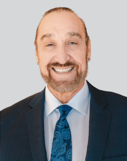 A man with a beard, smiling, wearing a suit and tie against a plain background.