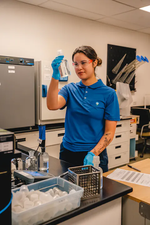 A person in a lab wearing gloves and goggles examines a bottle, with lab equipment around.
