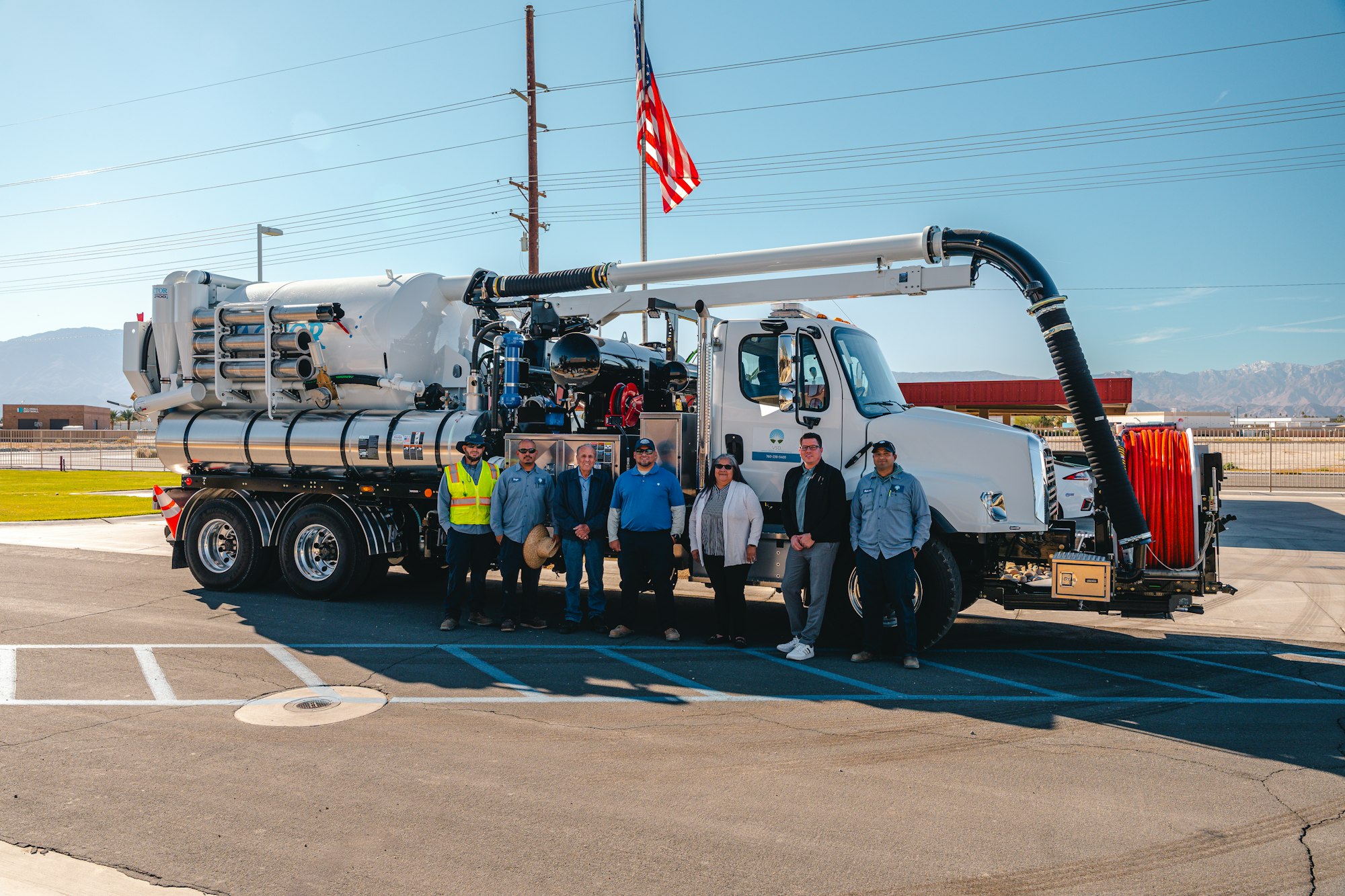 A group of people stands in front of a large service truck with equipment, an American flag in the background, under a clear sky.