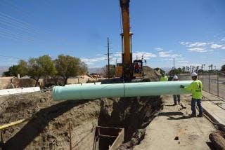 Construction workers use a crane to place a large pipe into a trench beside a road.