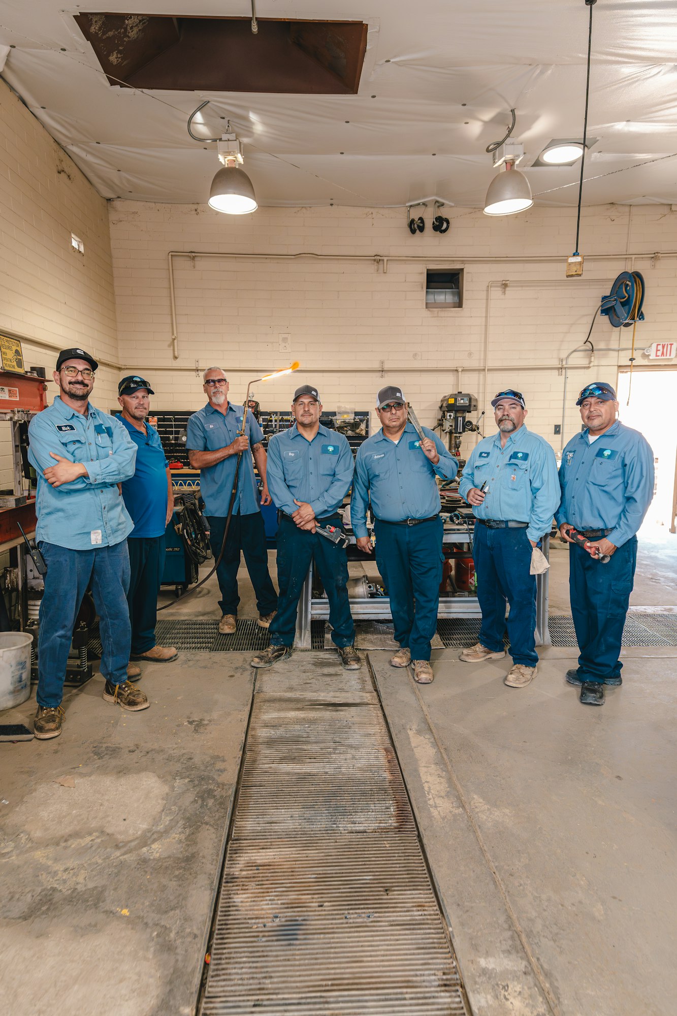 Seven men in matching blue work uniforms stand in a workshop. One holds a tool with a flame.