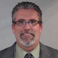 A person wearing a suit and tie, with glasses and a beard, smiling.