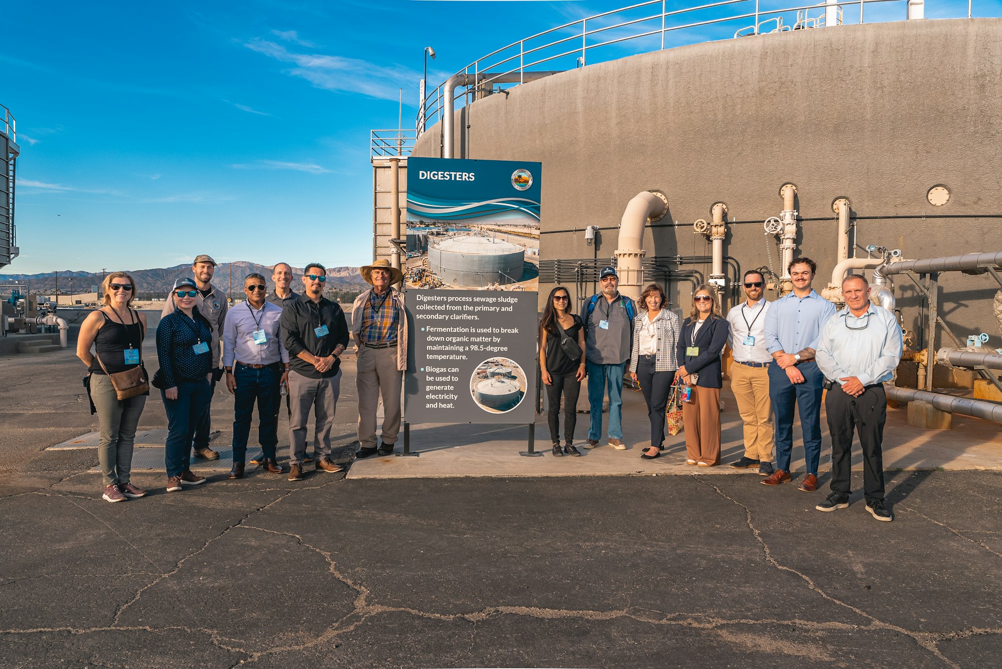 A group of people stand near a sign about digesters that process sewage sludge, with industrial tanks in the background.