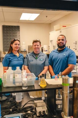 Three people in a lab setting, wearing blue uniforms, surrounded by various lab equipment and bottles.