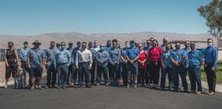 A group of people standing outdoors on a sunny day, wearing casual and uniform clothing, with mountains in the background.
