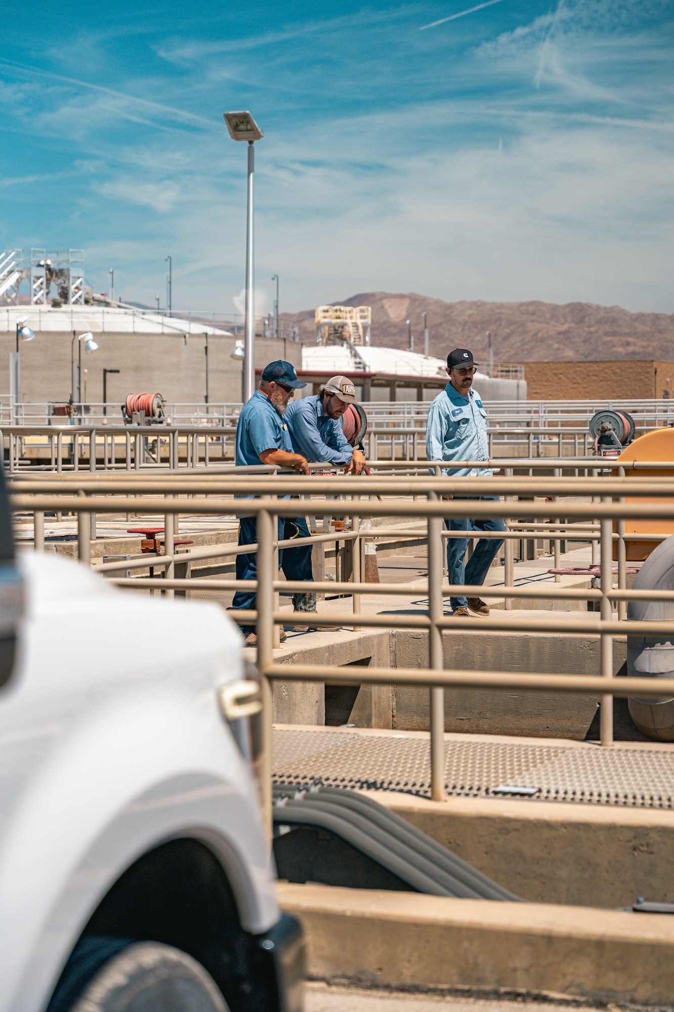 Three workers in blue uniforms standing on a platform, surrounded by industrial equipment, with mountains in the background.